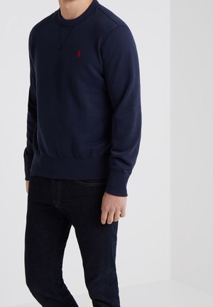 Uomo che indossa una felpa girocollo blu navy con logo rosso e jeans scuri, in piedi davanti a uno sfondo grigio chiaro uniforme.