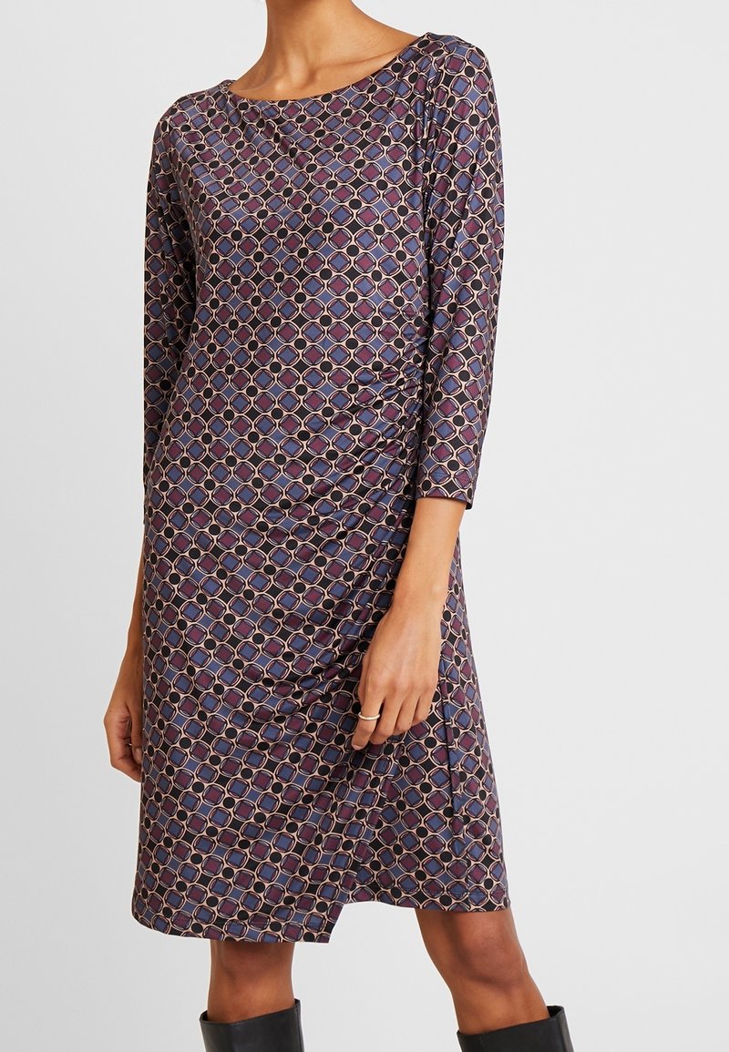 Femme portant une robe longue jusqu'aux genoux, manches longues, avec un motif géométrique aux tons violets, bleus et beiges, associée à des bottes noires.