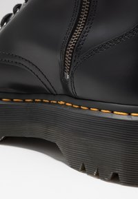 Gros plan d'une botte en cuir noir avec fermeture éclair latérale, couture jaune et semelle en caoutchouc épaisse à nervures avec motif de la bande de roulement.