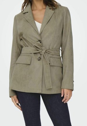 Kvinde iført en khakifarvet blazer med bælte, klaplommer, knaplukning og mørkeblå jeans, stående foran en ensfarvet hvid baggrund.