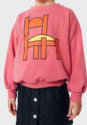 Child wearing a pink sweatshirt with an orange and yellow abstract chair design and a dark blue pinstriped skirt with white buttons.