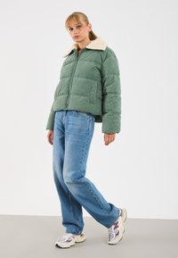 Woman wearing a green corduroy puffer jacket with shearling collar, blue jeans, and white sneakers standing against a plain background.