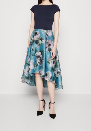 Femme portant une robe top manches courtes bleu marine, une jupe asymétrique bleue à motifs floraux avec un effet haut-bas, et des talons noirs à brides à la cheville.