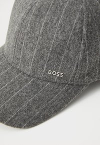 Grå uld baseballcap med lyse vertikale striber, prydet med et sølv "BOSS" logo foran, solid skygge og tekstureret stof.
