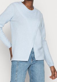 Pull en tricot bleu clair avec un col rond, des manches longues et une couture diagonale à l'avant avec des fentes latérales. Texture douce, coupe décontractée.