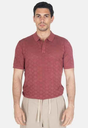 Uomo che indossa una polo bordeaux a maniche corte con motivo e pantaloni beige con coulisse, in piedi davanti a uno sfondo chiaro e uniforme.