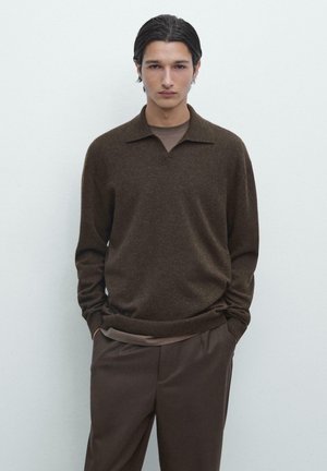 Giovane uomo con capelli scuri che indossa un maglione marrone con collo sopra una camicia marrone e pantaloni marroni, in piedi contro uno sfondo chiaro e uniforme.