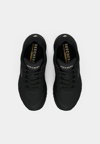 Čierne športové topánky s textúrovanými zvrškovými materiálmi, šnurovaním a polstrovanými vložkami. Obsahujú značku "SKECHERS" na jazyku a na boku.