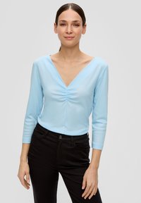 Hellblaues Longsleeve-Oberteil mit V-Ausschnitt und gerafftem Mittelpunkt, kombiniert mit schwarzen Hosen. Der Stoff erscheint weich und glatt.
