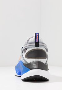 Grauer Sportschuh mit einem Mesh-Obermaterial, schwarzen und blauen Akzenten sowie einer strukturierten Gummisohle. An der Ferse befindet sich ein orangefarbenes Logoetikett.