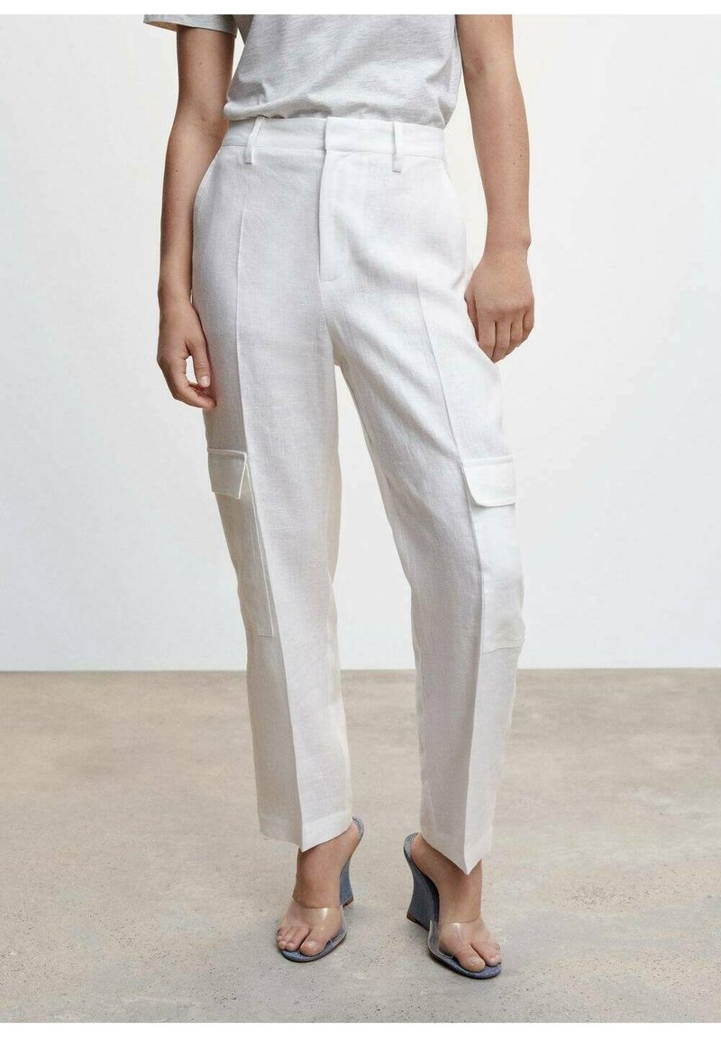 Personne portant un pantalon cargo blanc sur mesure et une chemise à manches courtes gris clair, debout sur un sol neutre avec des talons compensés à brides transparentes.