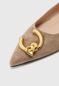 Scarpa beige in suede con punta a punta, caratterizzata da un marcato accento in metallo dorato in un design circolare e annodato sulla tomaia, con una fodera interna liscia.