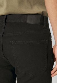 Vaqueros de mezclilla negra con un diseño ajustado, que presentan un parche de cuero en la cinturilla y un único bolsillo trasero con costuras sutiles.