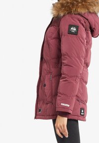 Chaqueta acolchada burdeos con capucha de piel, con textura acolchada, múltiples bolsillos y parches de marca en la manga y el pecho.