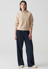 Felpa beige con cappuccio e tasca a marsupio, abbinata a pantaloni larghi blu scuro. Tessuto morbido, vestibilità rilassata e polsini a coste.