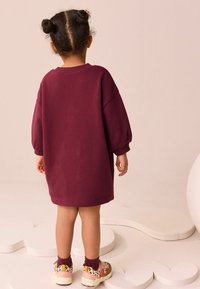 Burgunderfarbene Oversized-Sweatshirtkleid mit voluminösen Ärmeln. Ein Paar farbenfrohe, gesprenkelte Schuhe mit Knöchelsocken. Einfacher strukturierter Hintergrund.