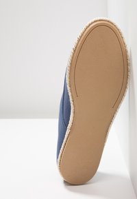 Chaussure bleue sans lacet avec semelle beige cousue, vue du dessous, appuyée contre une surface blanche.