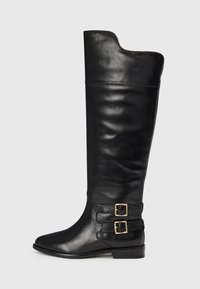 Botas de cuero negras hasta la rodilla con un acabado suave, punta afilada y dos hebillas en el lado exterior. Diseño de tacón plano.