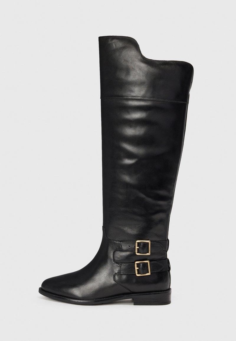 Botas de cuero negras hasta la rodilla con un acabado suave, punta afilada y dos hebillas en el lado exterior. Diseño de tacón plano.