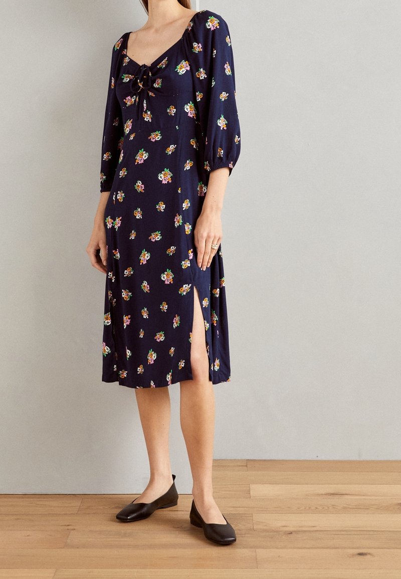 Femme portant une robe midi bleu marine à manches longues, imprimé floral, col à nouer devant, fente latérale, et chaussures plates noires.
