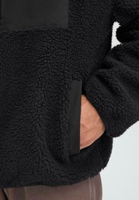 Hand in pocket of black textured fleece jacket with fabric trim, worn over brown pants.