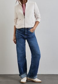 Kobieta w białej kurtce zapinanej na zamek, różowej koszuli, szerokich niebieskich dżinsach z mankietami i białych sneakersach, stojąca z jedną ręką w kieszeni.