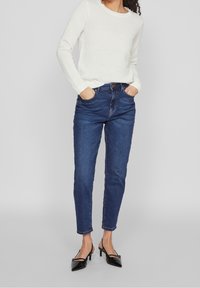 Pull en maille texturée blanche avec col rond, associé à un jean taille haute bleu foncé et des escarpins noirs à bout pointu.