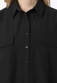 Chemise noire à manches longues avec col classique, deux poches poitrine et boutons noirs. Le tissu a une texture lisse.