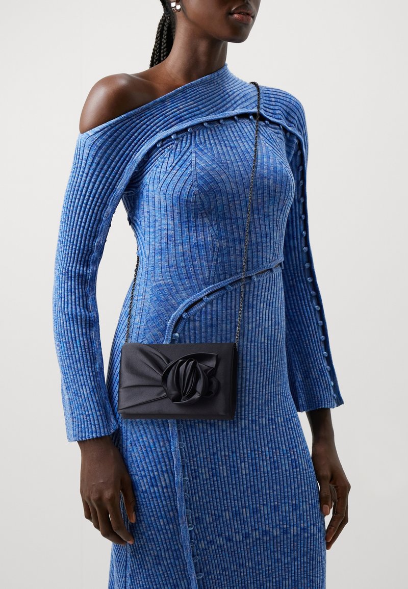 Femme noire portant une robe bleue côtelée à épaules dénudées avec des détails de boutons et une petite pochette noire avec un motif de fleur en tissu.