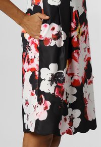Vestido floral de tela negra con grandes flores en tonos rosa y rojo, con un bolsillo lateral. El dobladillo tiene un borde con forma de concha para más detalle.