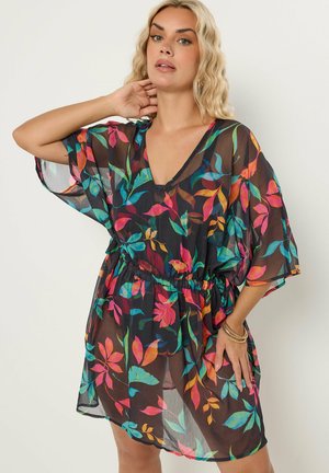 Frau trägt ein schwarzes, durchsichtiges Kleid mit bunten tropischen Blattmustern, weiten Ärmeln und tailliert, posiert mit einer Hand am Hals.