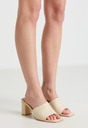 Gambe con mules beige a tacco largo e punta aperta, in piedi su una superficie chiara con sfondo neutro.