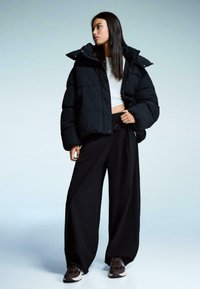 Zwarte pufferjack met een hoge kraag en oversized pasvorm, gecombineerd met brede zwarte broeken en donkere sneakers, tegen een gradient achtergrond.