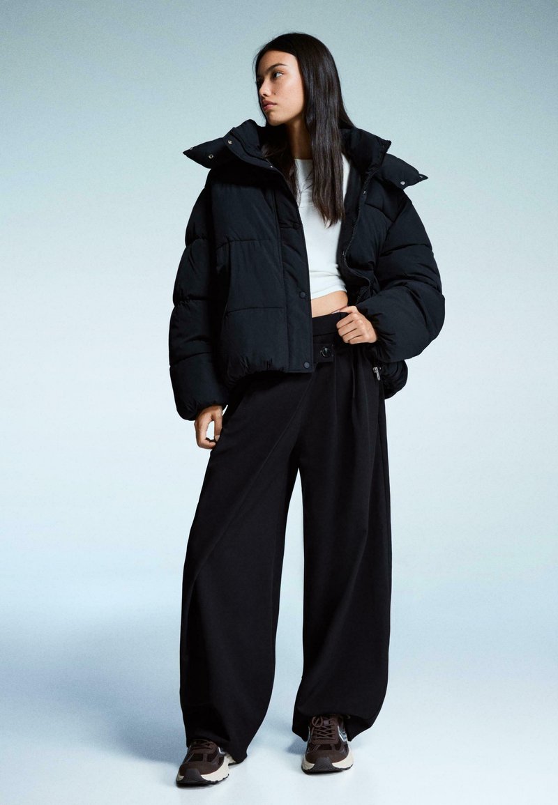 Zwarte pufferjack met een hoge kraag en oversized pasvorm, gecombineerd met brede zwarte broeken en donkere sneakers, tegen een gradient achtergrond.