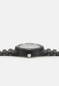 Montre numérique noire avec un bracelet en silicone texturé, un cadran rond et une couronne argentée. Présente de grands chiffres et une lunette multicolore.