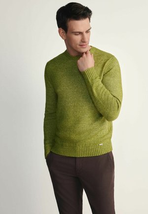 Zielony sweter z dzianiny z prążkowanymi mankietami i dołem, z okrągłym dekoltem i teksturowanym wykończeniem. Noszony z ciemnobrązowymi spodniami.