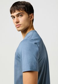 Camiseta de manga corta y cuello redondo en algodón azul claro con una textura suave. Presenta un discreto detalle de logo en el pecho.