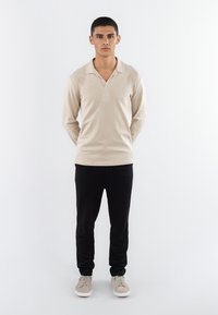 Beige Langarm-Polohemd mit Schalkragen, kombiniert mit schwarzen Jogginghosen. Das Modell trägt hellgraue Sneakers und steht vor einem weißen Hintergrund.