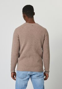 Bruine geribbelde trui met raglan mouwen, voorzien van een ronde halslijn en een kleine knoopdetail aan de achterkant. Draagt met lichtblauwe jeans.
