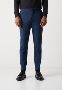 Blauwe textuurbroek met een elastische tailleband en een trekkoord, met een slim fit ontwerp, gedragen met zwarte instappers en sokken.