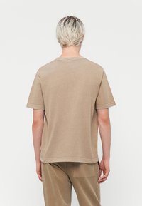 Khaki majica s kratkimi rokavi in okroglo ovratnikom, ki ima sproščen kroj in vidne šive. Narejena iz mehke bombažne tkanine.