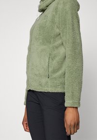 Groene fleece hoodie met een gestructureerd oppervlak, een ritszak aan de zijkant en een verstelbare trekkoordkap. Gecombineerd met zwarte broek.