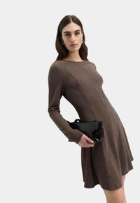 Brązowa sukienka z długim rękawem, z dopasowanym gorsetem, rozkloszowaną spódnicą i pionowymi szwami, w zestawieniu z czarną torebką typu kopertówka o fakturowanej powierzchni.