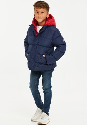 Marineblå pufferjakke med rød hætte, med stik-syninger og sidelommer. Bæres med blå jeans og hvide sneakers.