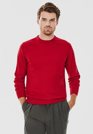 Czerwony sweter dziany z okrągłym dekoltem i długimi rękawami, w połączeniu z ciemnoszarymi spodniami. Materiał wydaje się gładki i dopasowany.