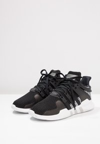 Svarta stickade sportskor med vita sulor, svarta snören och plaststöd på sidan, visade i en vinkel på en vit yta mot en ljus bakgrund.