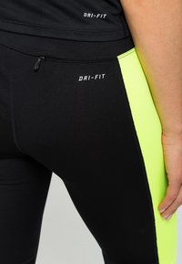 Czarne legginsy Dri-FIT z neonowo-żółtym bocznym panelem i małą zamykaną kieszonką. Gładka tekstura z logo umieszczonym blisko pasa.