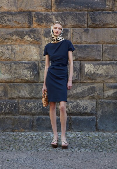 Abito fitted color navy con maniche corte, tessuto testurizzato e dettaglio torcigliato in vita. Abbinato a una sciarpa fantasia e una borsa intrecciata.