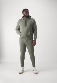 Conjunto de sudadera y pantalones jogger en verde oliva hecho de tejido texturizado. Presenta una capucha, bolsillo frontal y puños ajustados en los tobillos. Zapatillas con detalles.