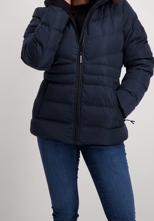 Osoba ubrana w granatową pikowaną kurtkę z przednim suwakiem i bocznymi kieszeniami, w połączeniu z niebieskimi dżinsami na jednolitym tle.